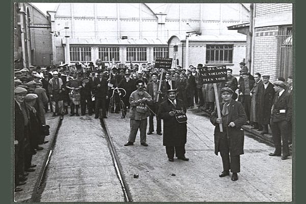 En France, même son de cloche: Front populaire, 1936, déguisements et sketches lors des grèves dans une cour d’usine. Un homme tient un panneau « Ce que nous ne voulons plus voir », et un homme, « Le capital », tient en laisse un camarade, « La misère » © DR – Mémoires d’Humanité / Archives départementales de la Seine-Saint-Denis